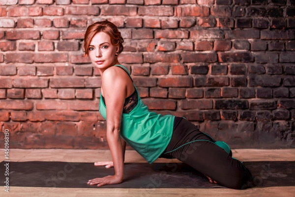 Fototapeta Young slender athletic girl in a turquoise tank top and dark leggings doing yoga against the background of a brick wall