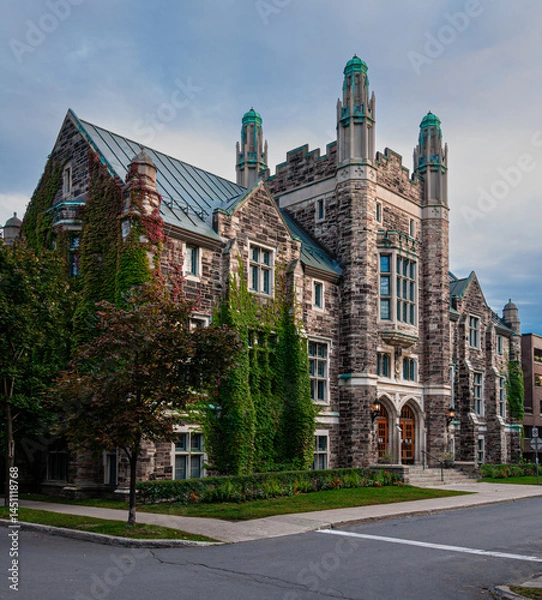 Obraz Elegant historic stone building with ivy-covered walls and green towers, photographed on a quiet street at sunset. Beautiful example of Gothic Revival architecture in an urban setting.