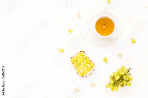 Fototapeta Healthy fruit breakfast. Cup of green tea, fruit toasts with grapes, cream cheese, berry, cereals and nuts on white background. Top view, flat lay.
