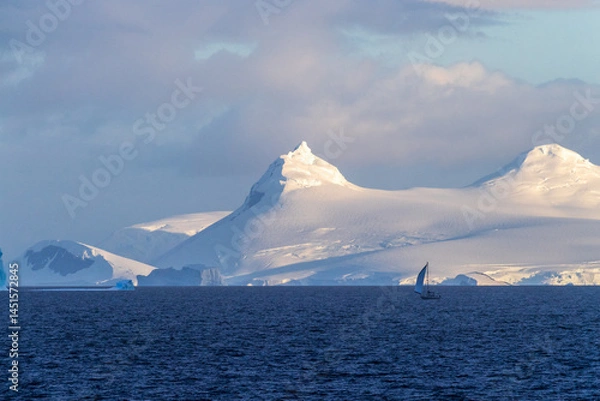 Fototapeta An Antarctic landscape, taken during the golden hour, just after sunrise, in the Gerlache Strait. A little sailboat in the foreground explores the Antarctic waters.