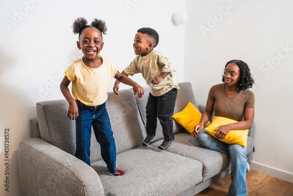 Fototapeta African two cheerful children are jumping on a grey sofa while their mother is smiling and watching them in their modern living room