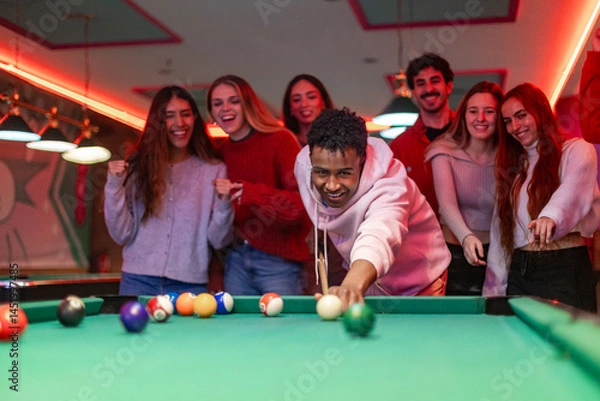 Obraz Young man playing billiard with friends cheering him up in a pub