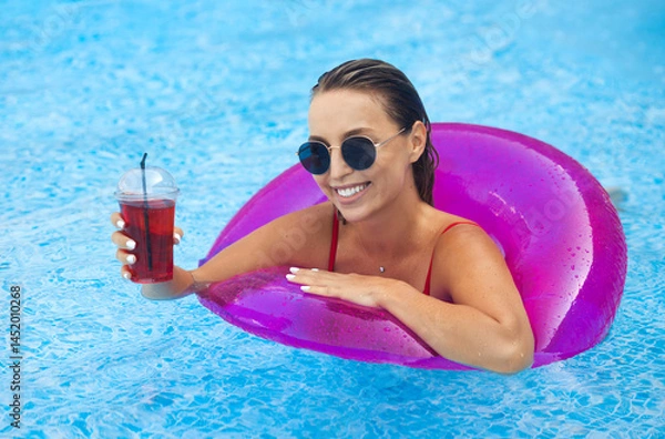 Fototapeta Gorgeous blonde model girl with a beautiful white smile swims in an inflatable rubber ring and drinks a cocktail. Happy woman in sunglasses relax at a summer resort in the swimming pool.