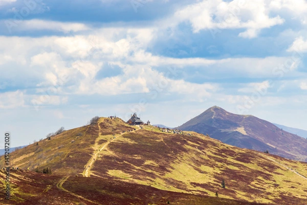 Obraz Beautiful mountain landscape showcasing a hiking trail leading to a peak with a small cabin, surrounded by lush hills under a blue sky with fluffy clouds.