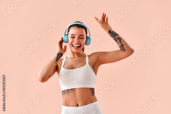 Fototapeta Stylish young woman with arm tattoos, wearing a white sports top and leggings, dancing with blue headphones, enjoying a fitness workout and maintaining a positive mood, against a soft pink background.