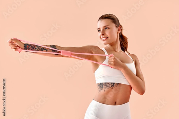 Fototapeta A sporty fitness girl, a trainer, is stretching her arms using a pink resistance band against a light background in a studio.