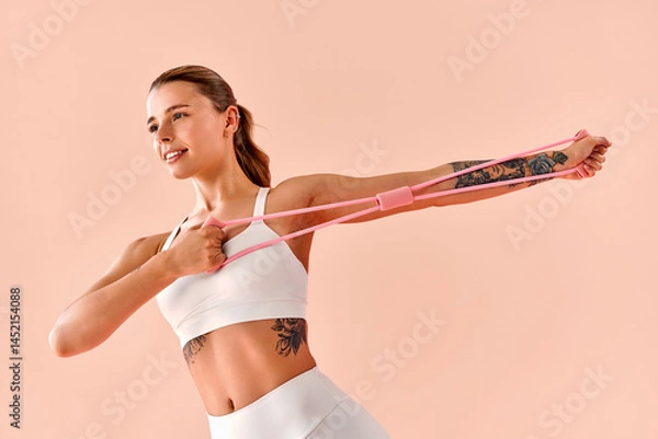 Fototapeta A toned athletic woman is stretching her arms using a pink resistance band against a soft pink background.