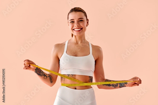 Fototapeta Happy, athletic, toned, modern young woman with tattoos on her arms is measuring her waist with a yellow tape measure and smiling against a light background.