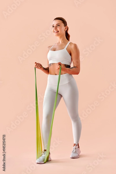 Fototapeta Vertical image – A fit young woman in white sportswear working out with a green resistance band against a peach background. Fitness, motivation, healthy lifestyle.