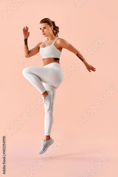Fototapeta A vertical full-body image of a sporty, toned young girl in white fitness wear, captured mid-jump against a soft pink background.