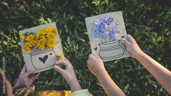 Obraz Two children creating flower crafts with fresh dandelions on cardboard, enjoying springtime nature and creative outdoor activity in a grassy field