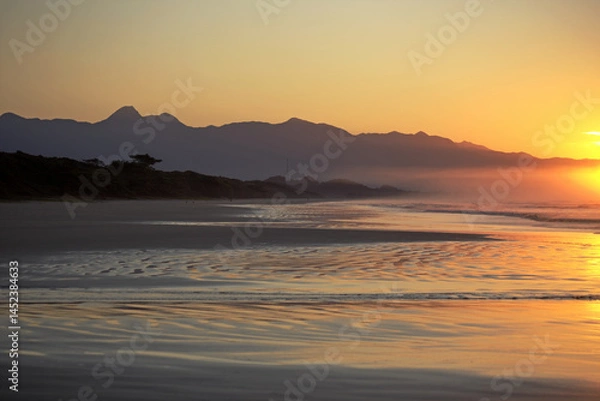 Obraz Sunrise in Itaguare beach, Bertioga, Brazil. Yellow light on sky and reflections on water, shadows over vegetation in silhouette and  mountains in background