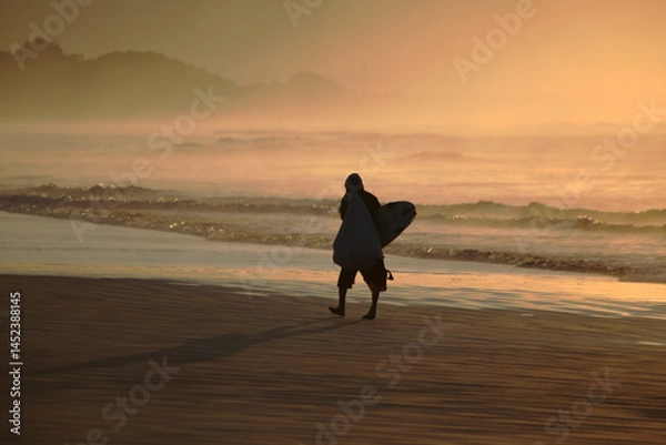 Obraz Surfer silhouette walking on the beach sand early morning before sunrise. Light reflections on sky and water. Itaguare beach, Bertioga, Brazil