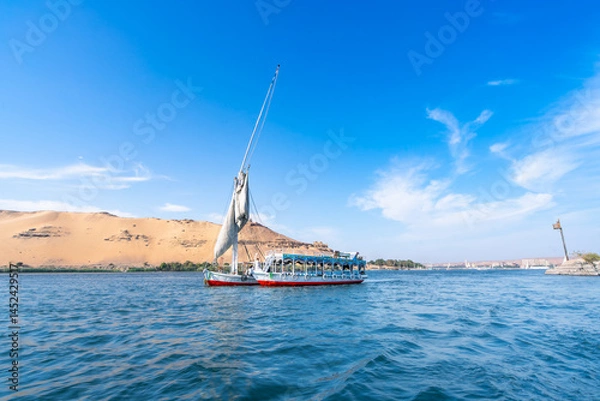 Obraz A felucca sailboat carrying tourists cruises along the Nile River near Aswan High Dam, Egypt, on a bright spring day with clear blue skies and light clouds.