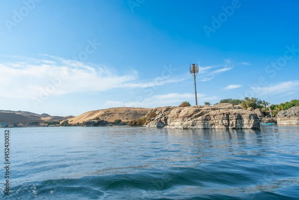 Obraz Aswan High Dam, Egypt: A serene Nile River view in spring, with rocky shores, greenery, and sandy hills under a clear blue sky.