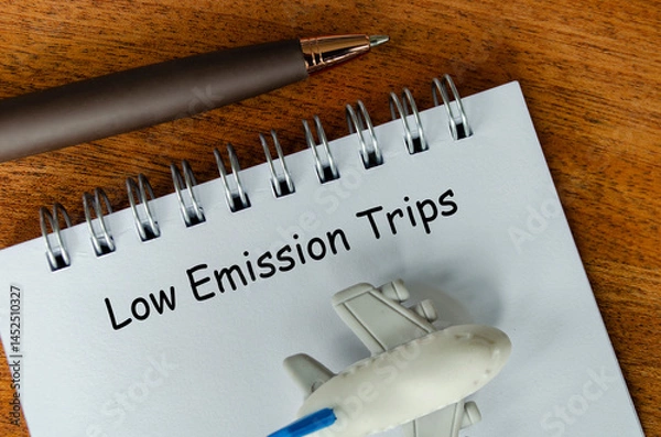 Fototapeta Top view of Low Emission Trips text on white notepad with small airplane and pen background. Environmental awareness