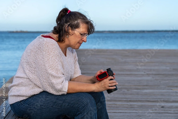 Fototapeta A woman is sitting on a bench by the water, holding a camera. She is enjoying the view and taking a photo in blue hour
