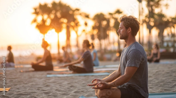 Fototapeta Young caucasian male practicing yoga on beach at sunset with diverse group in background