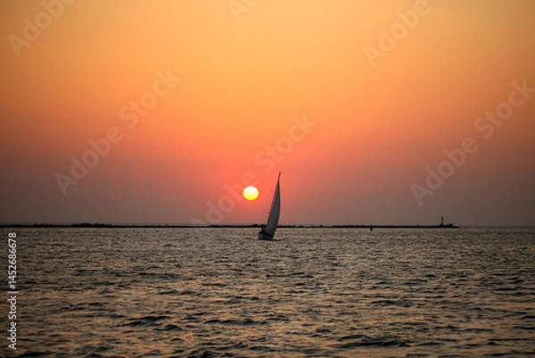 Obraz Sailboat at sunset over calm sea horizon Silhouette of a sailboat gliding across the ocean at sunset with a glowing orange sky