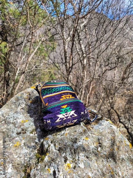 Obraz Bright small backpack with ethnic pattern on a rock, shot in close-up. Blurred background with dry forest in mountains, spring sunlight. Natural colors, travel concept, outdoor lifestyle.