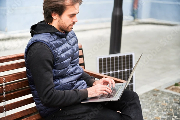 Obraz Man sits on wooden bench, working on laptop connected to solar panel. Urban setting and casual attire highlight modern, sustainable, mobile, eco-friendly workspace.