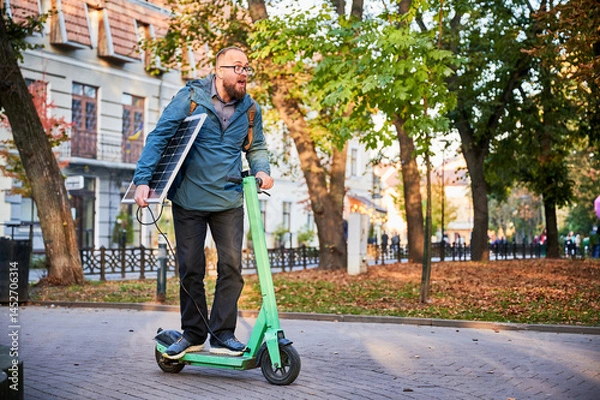 Obraz Man rides electric scooter while carrying solar panel. Integration of solar power as sustainable energy source for charging electric scooters, promoting eco-friendly urban transportation.