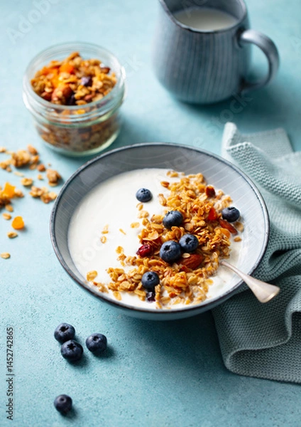 Fototapeta Delicious bowl of yogurt topped with crunchy granola and ripe blueberries. A jar of granola and a jug of milk are in the background.