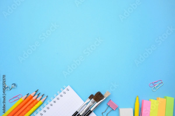 Fototapeta Colorful stationery, brushes, and notebooks arranged on pastel blue background. Ideal for school banners, creative ads, and online courses.