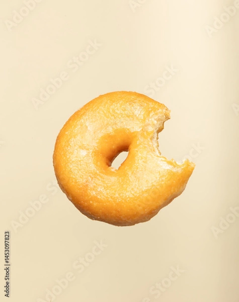 Fototapeta Bitten glazed donut floating on a soft beige background. Minimal and creative food concept ideal for bakery, dessert, or snack-related designs and modern visual projects.