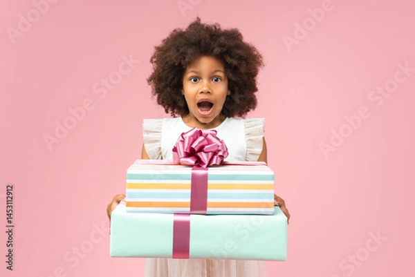 Fototapeta Light colored portrait of excited African American birthday girl posing in studio with a pile of gift boxes in hands with surprised open mouth face expression, isolated on pastel pink background