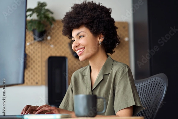 Fototapeta Happy smiling young busy African American woman student or employee using pc typing elearning or hybrid working at home office looking at computer learning, studying online sitting at work desk.