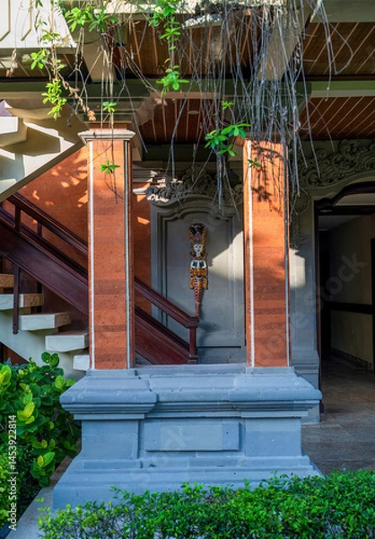 Obraz Traditional Balinese entryway or home entrance with red columns, ornate woodwork, hanging plants, and a decorative deity figure mounted on the wall.