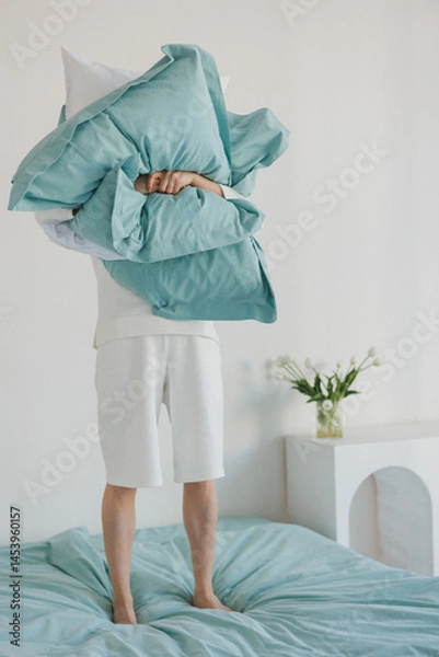 Obraz A person is currently standing on a bed while holding a fluffy pillow high above their head, creating a playful atmosphere in the room