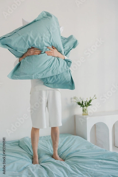 Obraz A person is holding a large, soft pillow high above their head while standing on a cozy bed that is covered with colorful linens