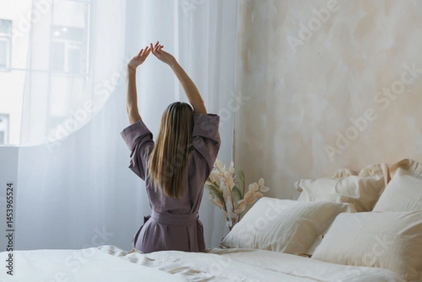 Obraz A woman is comfortably sitting on a soft bed stretching her arms in front of a large window, letting in natural light through it