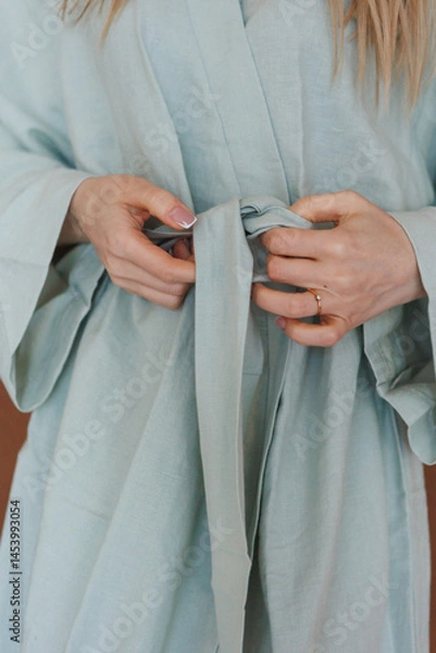 Obraz A woman dressed in a light blue robe is carefully tying a belt around her waist, showcasing the beauty of her attire