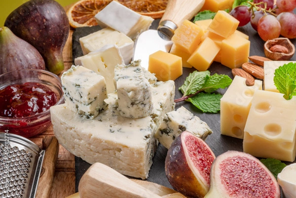 Fototapeta Variety of sliced cheeses with fruits, mint, nuts and cheese cutting knives.