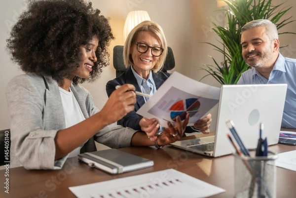 Fototapeta Three busy happy middle aged professional business man and two women executive leaders team using laptop working on computer at work desk having conversation on financial project at meeting in office.