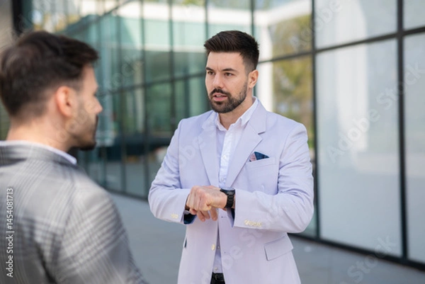 Fototapeta Serious businessman explaining something while checking his watch during an outdoor conversation with a colleague.