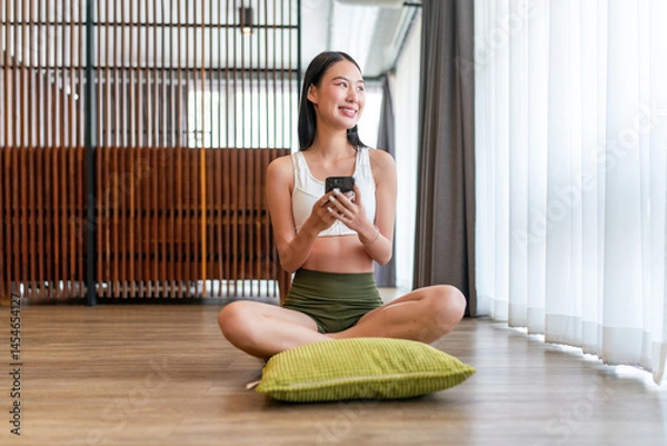Obraz Asian Woman Sitting Cross-Legged at Home Using Phone. Young Lady Smiling While Enjoying a Peaceful Digital Moment Indoors.