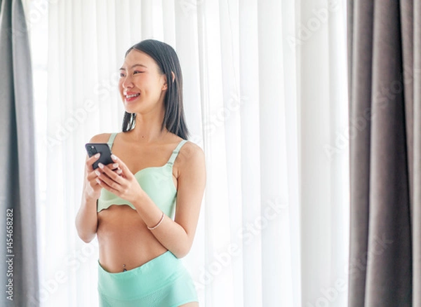 Fototapeta Asian Woman Using Phone by Window in Underwear. Young Lady Smiling While Enjoying a Relaxed Digital Morning at Home.