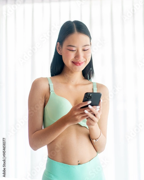 Fototapeta Asian Woman Using Phone by Window in Underwear. Young Lady Smiling While Enjoying a Relaxed Digital Morning at Home.