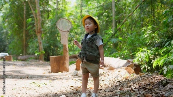 Obraz A Young Explorer Asian child dressed for adventure with Hat stands in a forest holding a net with butterflies, ready to explore Discovering the wonders of nature in Lush Greenery