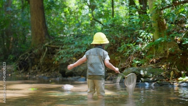 Obraz A young Asian child wearing a yellow hat and vest explores a forest stream with a fishing net, surrounded by lush greenery on a Sunny Day, ready to Discovering the wonders of nature