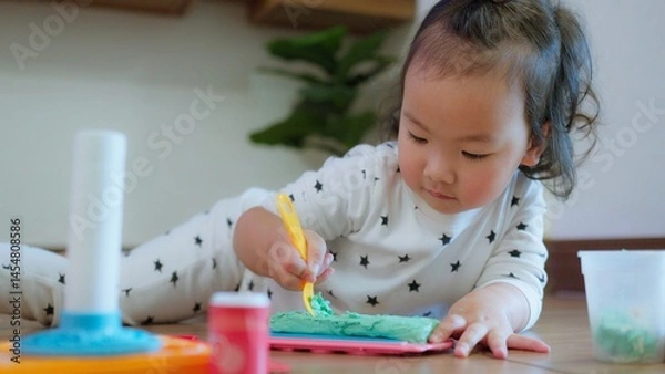 Obraz Cute Asian toddler child girl playing with modeling clay on Wooden Floor, enhancing creativity education and motor skills in a cozy home environment.