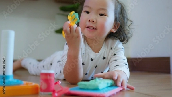 Obraz Cute Asian toddler child girl playing with modeling clay on Wooden Floor, enhancing creativity education and motor skills in a cozy home environment.