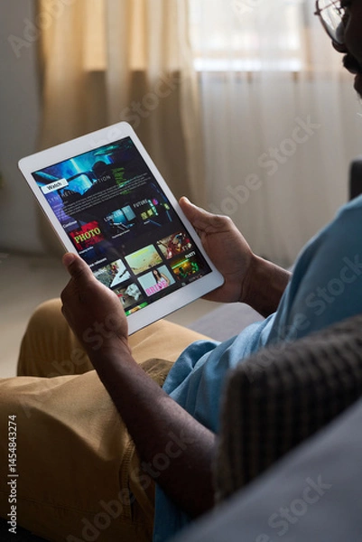 Fototapeta Vertical medium shot of African American man sitting on couch at home, watching movie or TV show trailer on streaming app on tablet