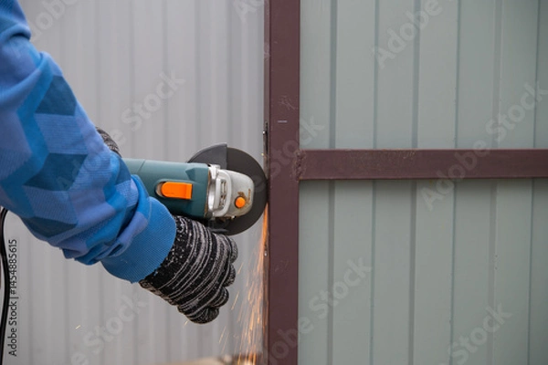 Fototapeta A man cuts metal with an angle grinder, causing a lot of sparks. An angle grinder is a handheld power tool used for grinding (abrasive cutting) and polishing.