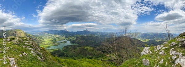 Fototapeta Breathtaking panoramic view embracing lush green valley, serene lake and majestic mountains under cloudy sky.