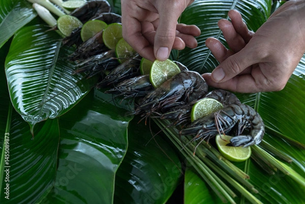 Fototapeta Hands skillfully place fresh tiger shrimp atop vibrant banana leaves, garnished with lime slices, creating an inviting display for a tropical feast.